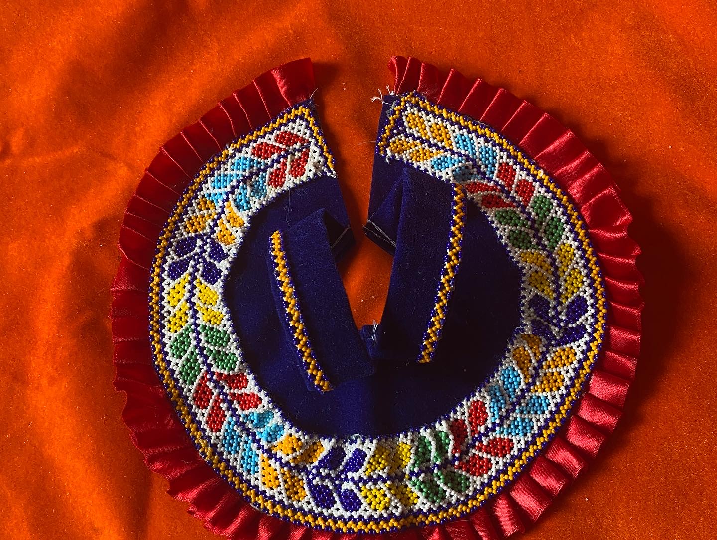 Circular Laddu Gopal aasan with multicoloured beadwork forming leaf patterns on dark blue fabric with red satin ruffle